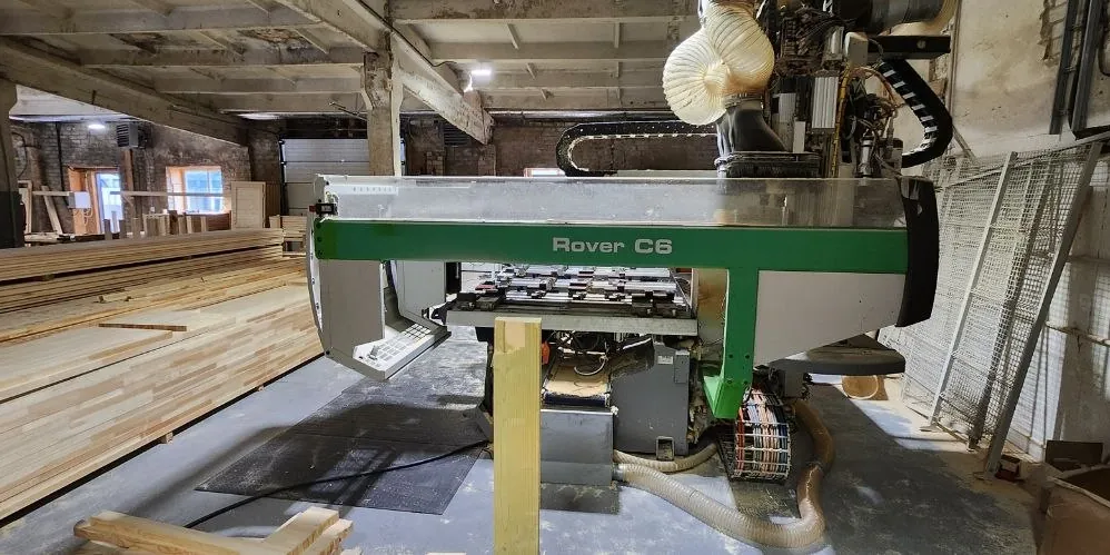 A CNC machining centre in a brick-walled workshop, with stacks of timber blanks on the left