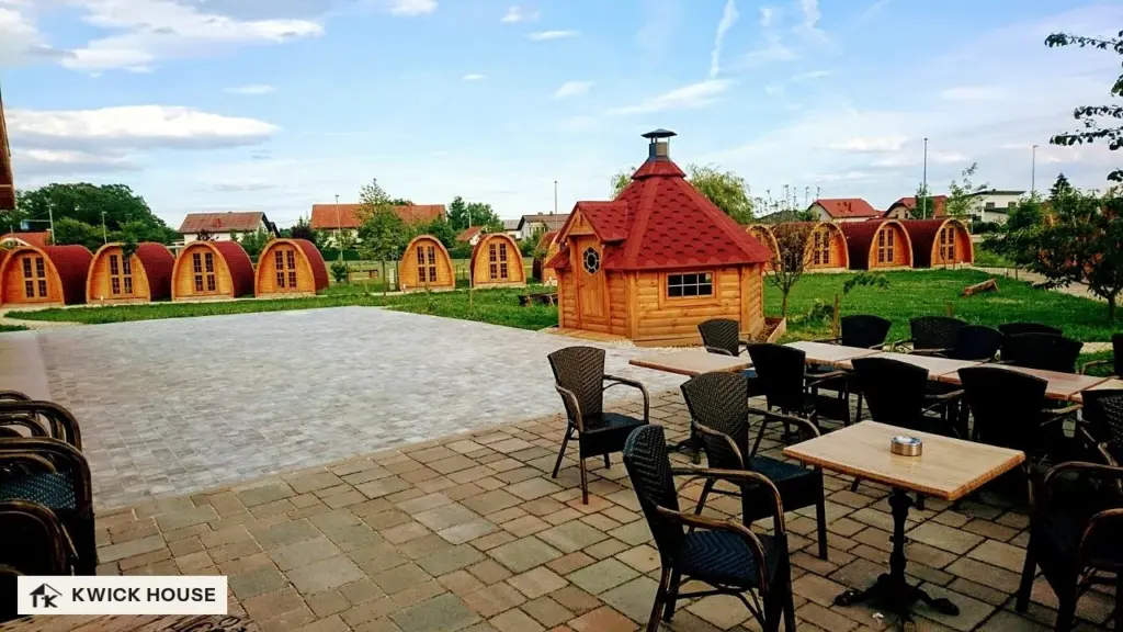 Row of arched glamping pods with red roofs around a lawn, seen from a paved terrace with tables and chairs