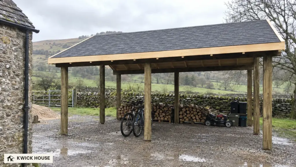 Wooden Carport 36 m², Apex Roof, seen from the long side on a wet gravel farmyard, with stone walls and hills beyond